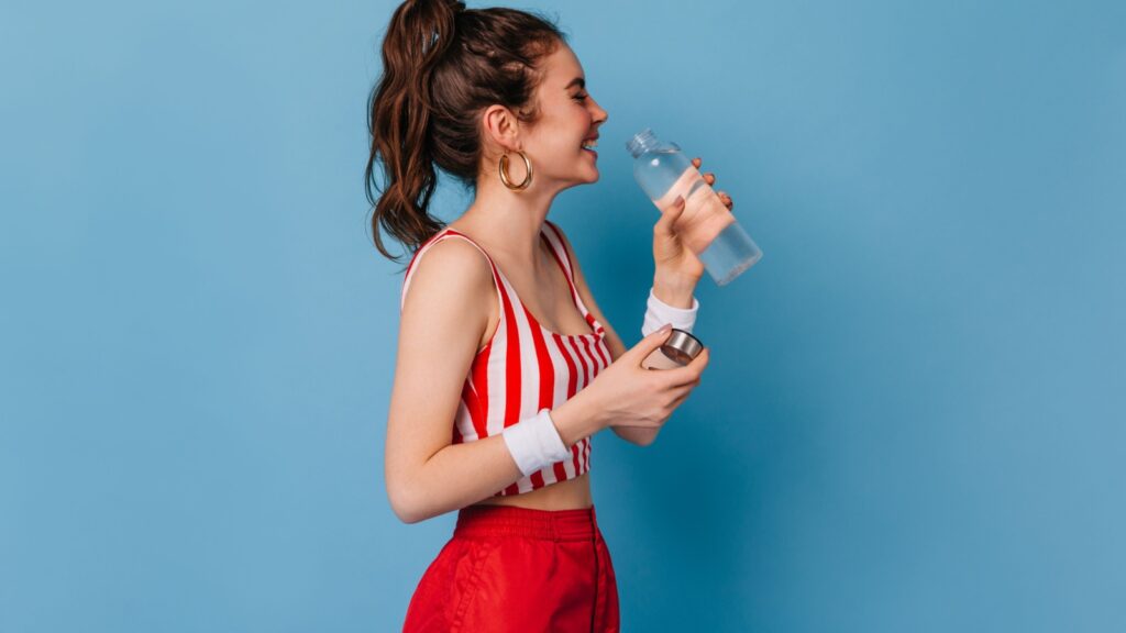 Young girl in red striped outfit laughs and drinks water from bottle on isolated background