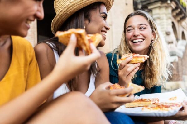 Three young multiracial women laughing while eating a piece of pizza in italian city street - Happy female friends enjoying holidays together in Italy - Friendship, travel and tourism concept