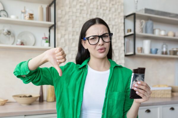 Woman at home disapproving of a bar of chocolate, looking at the camera and pointing a finger down, brunette at home in the kitchen in a green shirt and glasses