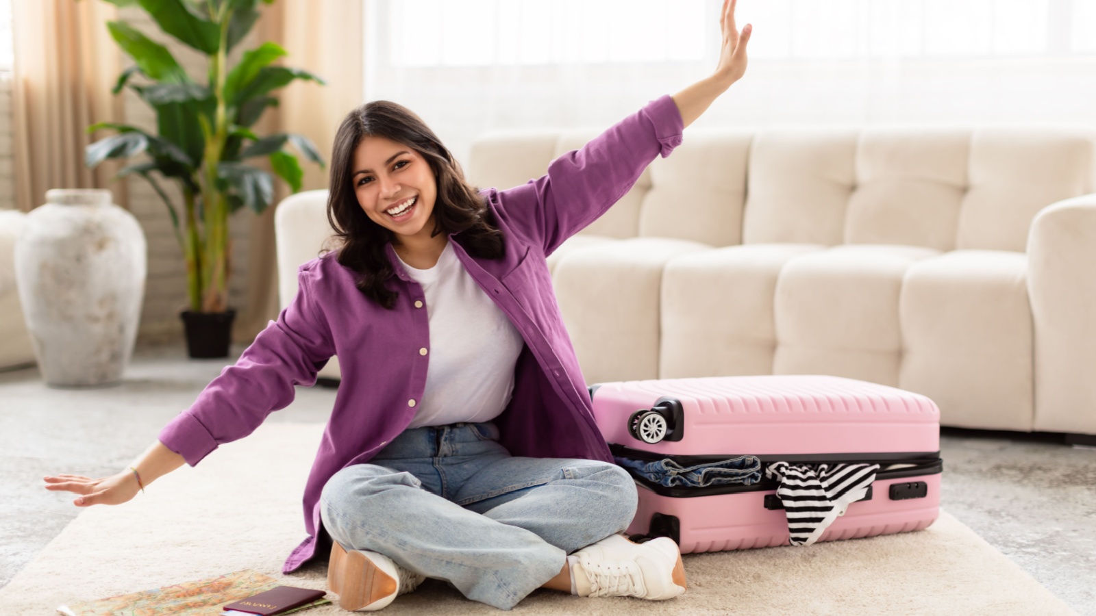 A joyous middle eastern woman with arms wide open sitting on the floor next to a pink suitcase, expressing travel excitement