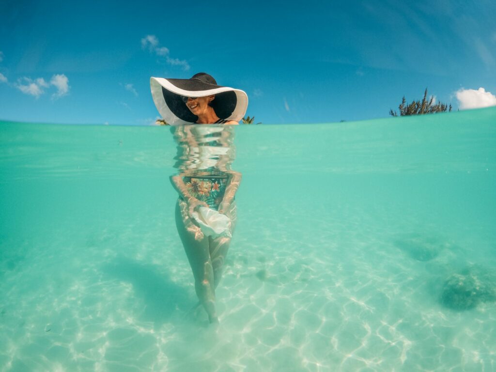 A woman wearing a large sunhat and colorful one piece swimsuit standing in clear blue water holding a conch shell, the beach is in the background