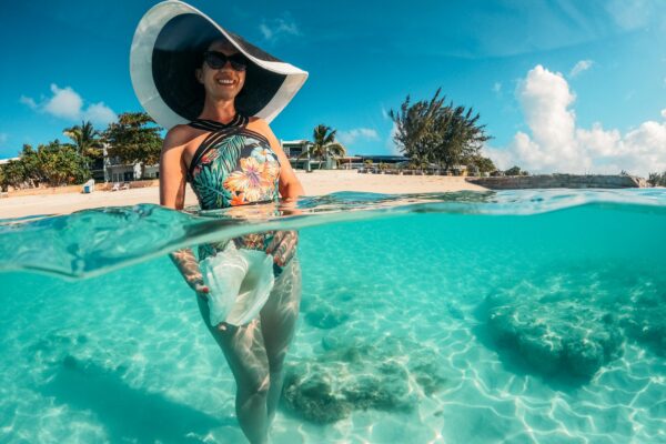 A woman wearing a large sunhat and colorful one piece swimsuit standing in clear blue water holding a conch shell, the beach is in the background