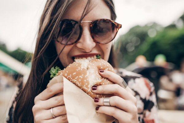 stylish hipster woman eating juicy burger. boho girl biting yummy cheeseburger, smiling at street food festival. summertime. summer vacation travel picnic.