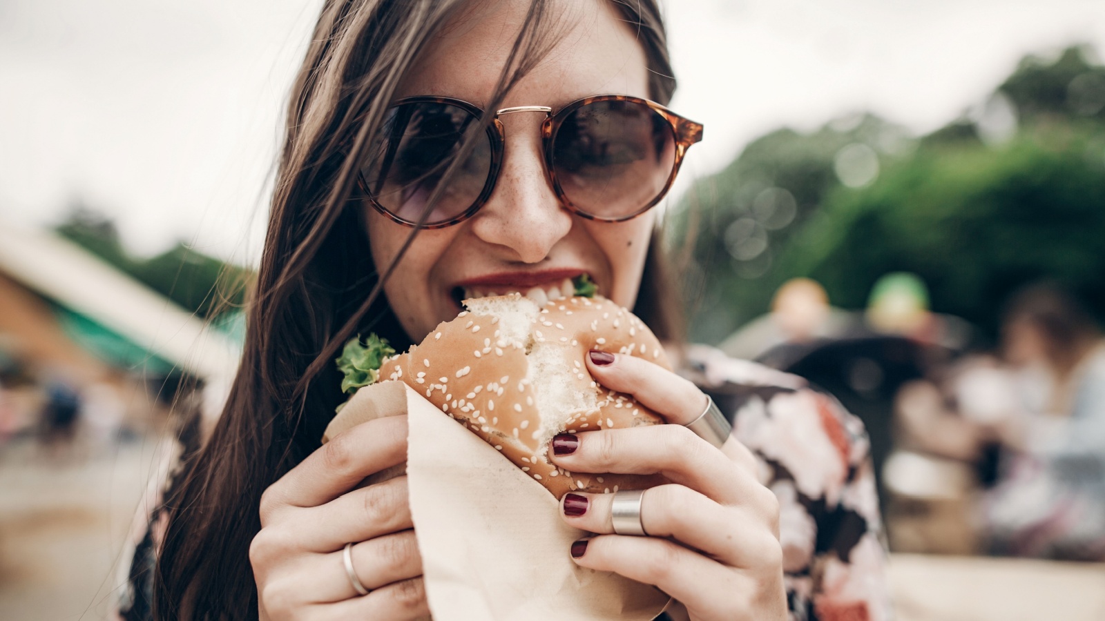 stylish hipster woman eating juicy burger. boho girl biting yummy cheeseburger, smiling at street food festival. summertime. summer vacation travel picnic.