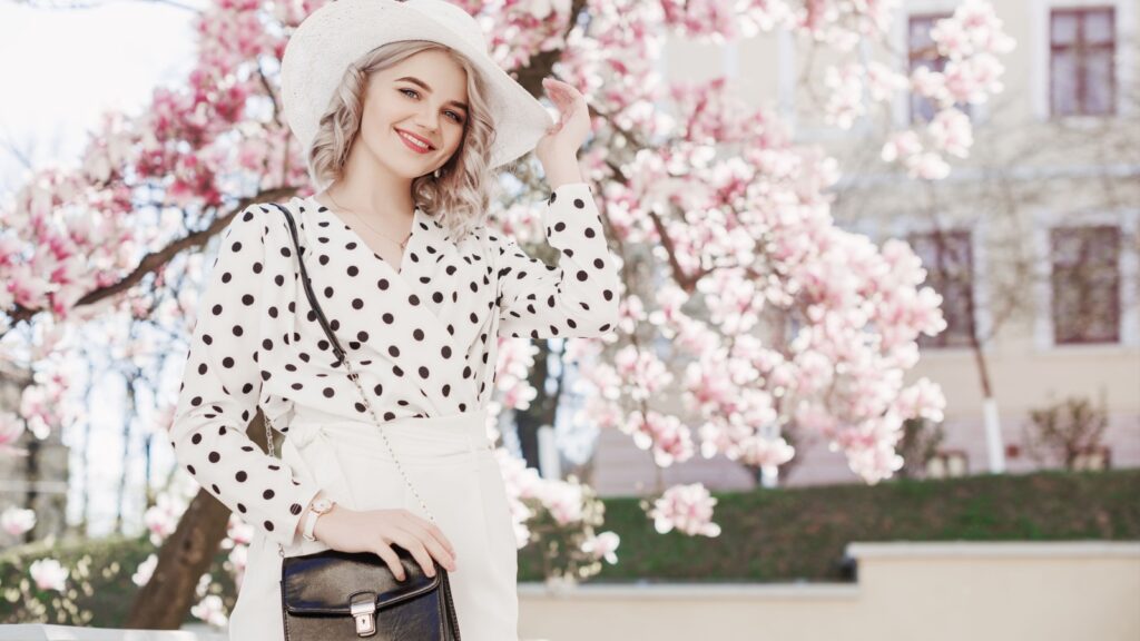 Outdoor fashion portrait of young beautiful happy smiling lady wearing stylish white hat, wrist watch, polka dot blouse, holding small bag, posing in street with blooming magnolia tree. Copy space