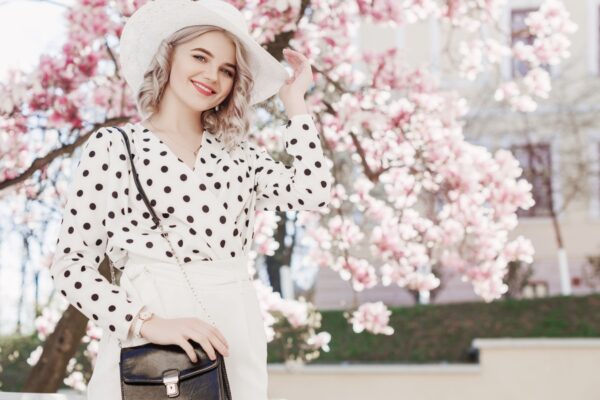 Outdoor fashion portrait of young beautiful happy smiling lady wearing stylish white hat, wrist watch, polka dot blouse, holding small bag, posing in street with blooming magnolia tree. Copy space