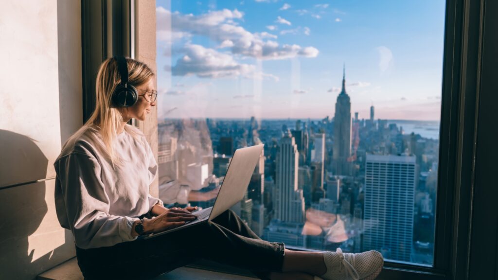 Millennial female freelancer in electronic headphones for noise cancellation listening positive music podcast and looking in panoramic window with breathtaking Manhattan view from high rise