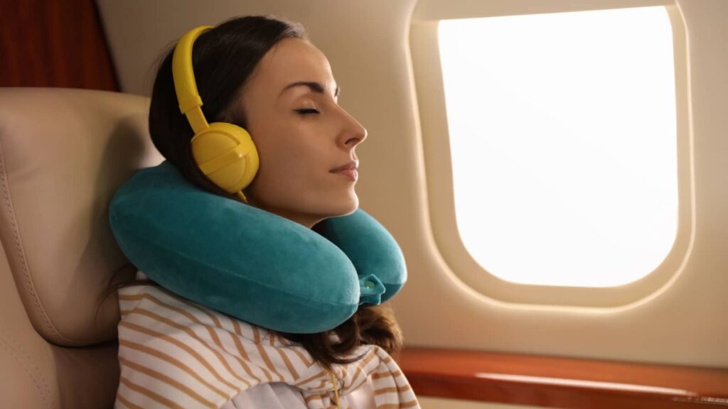 Young woman with travel pillow resting while listening to music in airplane during flight