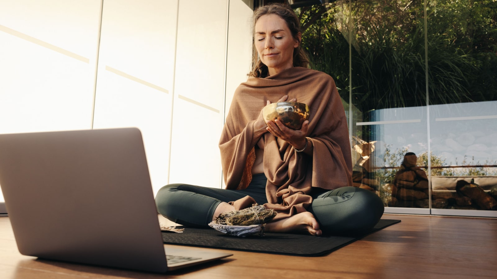 Ayurvedic healer meditating with a singing bowl and sage during an online holistic class. Woman performing a self-healing and purifying ceremony at home. Senior woman taking care of her ageing body.
