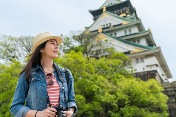 full length of young girl travel photographer walking along the bamboo walls of japanese style wooden house with trees and graden inside. woman backpacker on street road in town city kyoto japan.