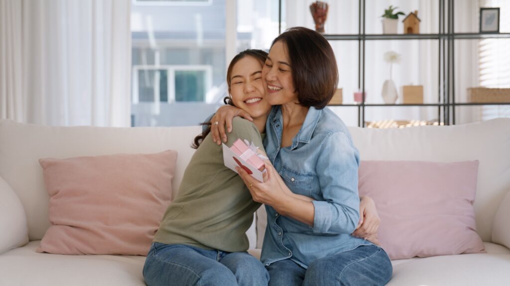 May Mother's day young adult grown up child cuddle hug give flower gift box red heart card to mature middle aged mum. Love kiss care mom asia people sitting at home sofa happy smile enjoy family time.