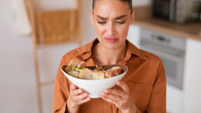 Young dissatisfied european woman eating caesar salad looking at it, dont like it smell and feeling disgust while sitting in kitchen interior, above view