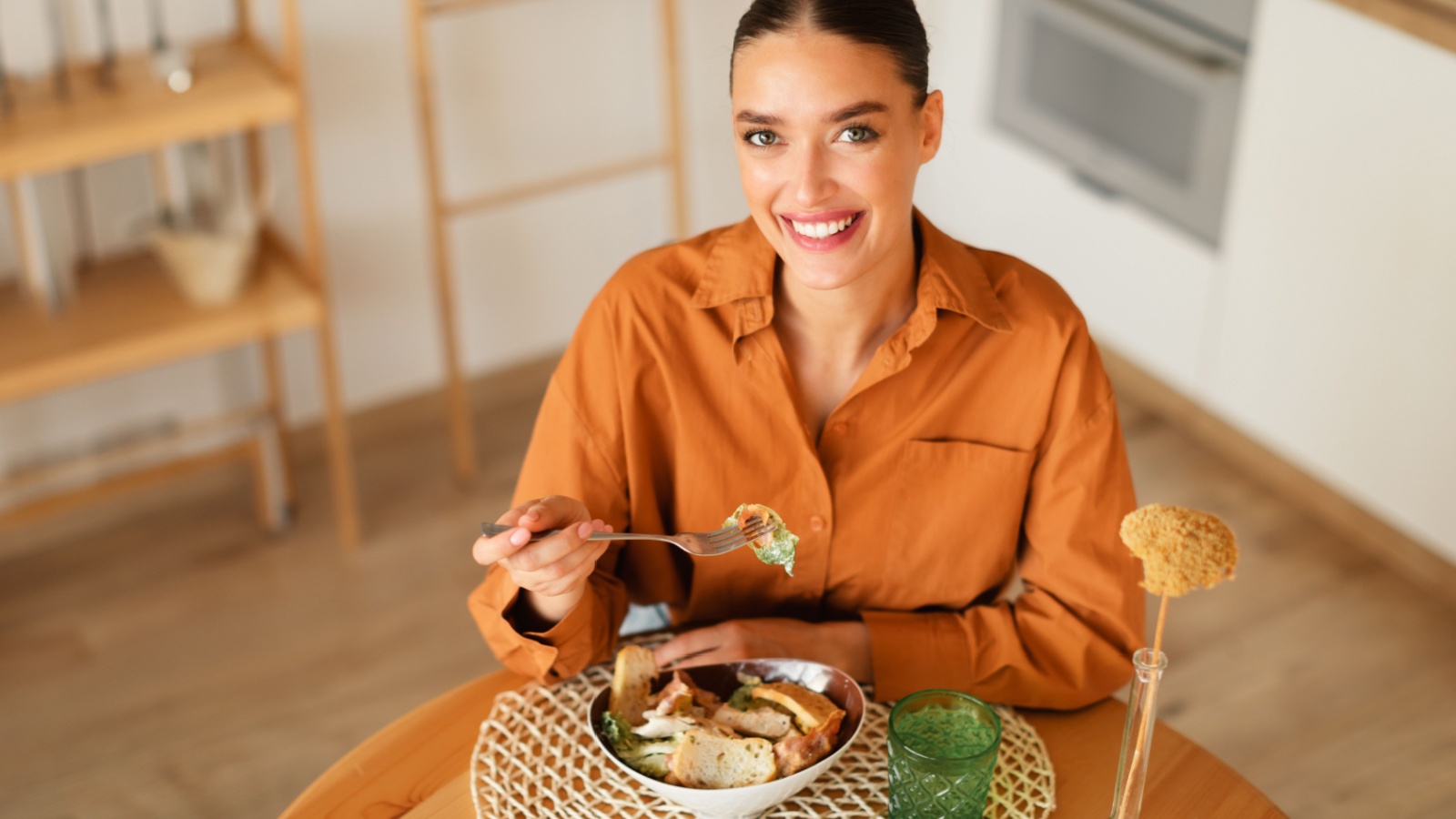 Happy young european woman enjoying freshly made caesar salad, sitting at table in kitchen interior, looking and smiling at camera, copy space, above