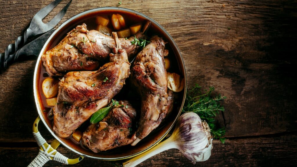 High Angle View of Roasted Rabbit Haunches in Pan with Stewed Vegetables on Rustic Wooden Table Surface with Bulb of Garlic and Copy Space