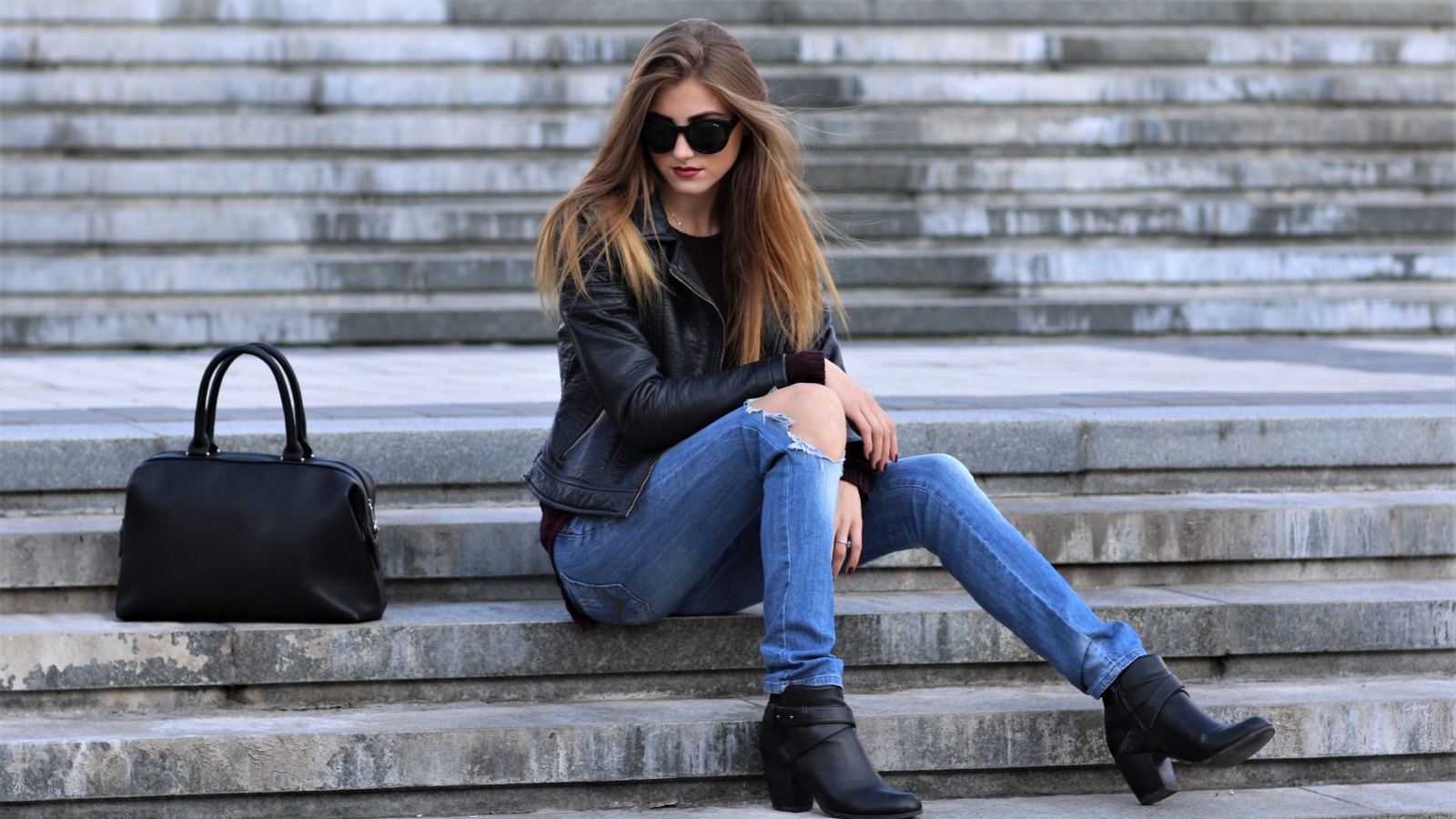 Casual style. Stylish woman in sunglasses sitting on the stone stairs. She dressed in leather jacket, skinny jeans and shoes on heels. Her sight directed downwards. Long hair, elegant, street fashion