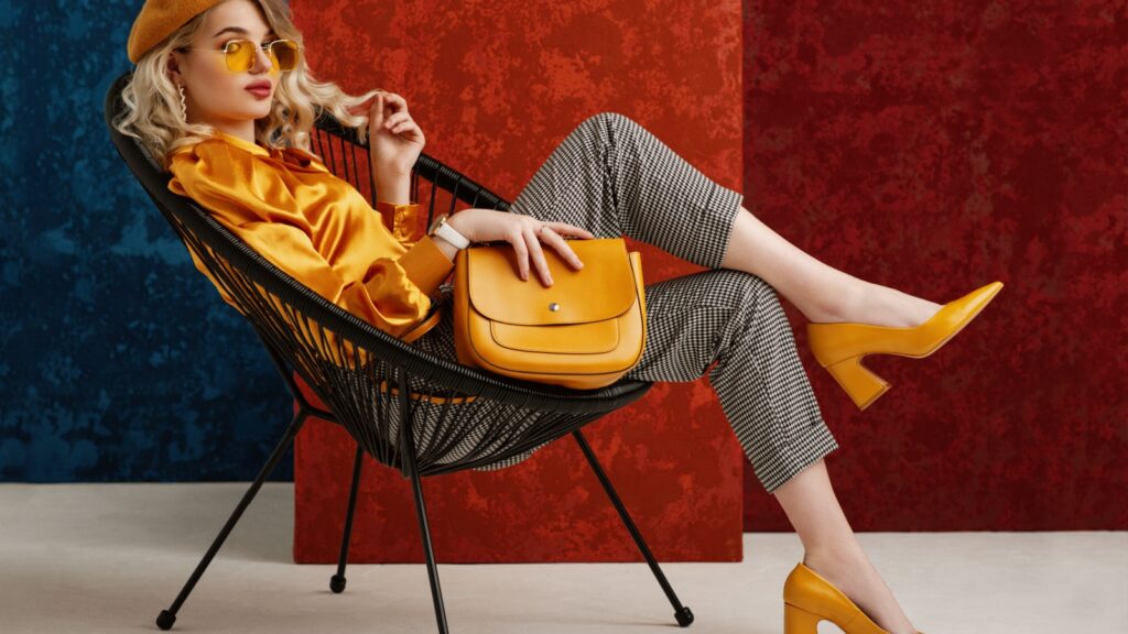 Full-length studio fashion portrait of elegant woman wearing yellow color sunglasses, beret, silk blouse, houndstooth printed trousers, pointed toe shoes, posing on chair, holding stylish leather bag