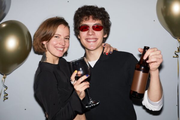 Waist up portrait of smiling young couple partying and looking at camera holding drinks, shot with flash