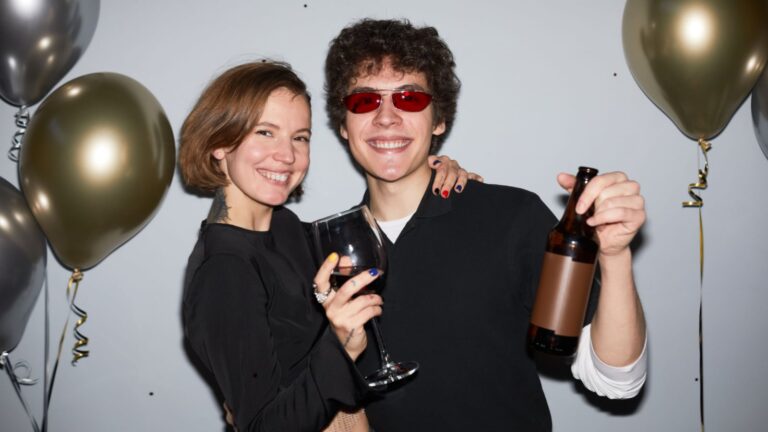Waist up portrait of smiling young couple partying and looking at camera holding drinks, shot with flash