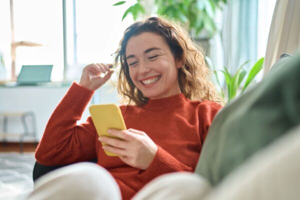 Happy relaxed young woman sitting on couch using cell phone, smiling lady laughing holding smartphone, looking at cellphone enjoying doing online ecommerce shopping in mobile apps or watching