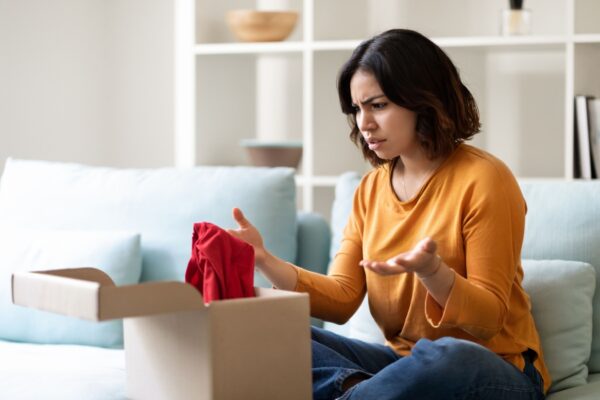 Wrong Parcel. Confused arab woman looking at open cardboard box at home and frowning, shocked middle eastern female disappointed about shipping mistake, feeling angry because of bad delivery service