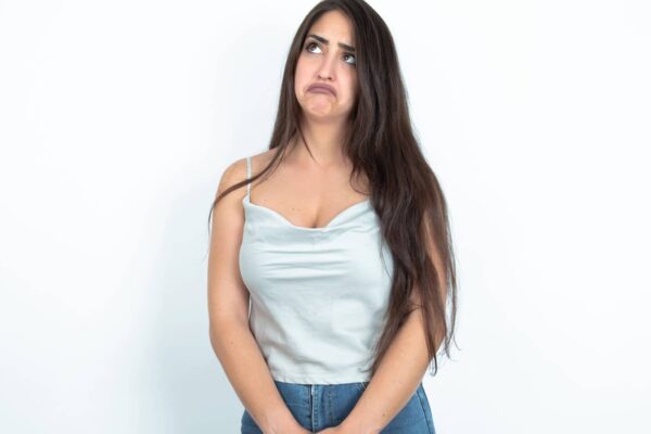 Dissatisfied young brunette woman wearing white tank top over white studio background purses lips and has unhappy expression looks away stands offended. Depressed frustrated model.