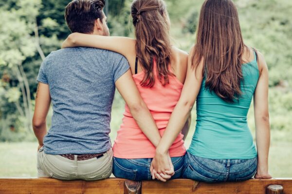 Love triangle, a girl is hugging a guy and he is holding hands with another girl, they are sitting together on a bench