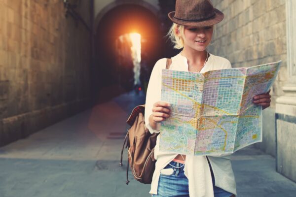 Cheerful woman wanderer with trendy look searching direction on location map while traveling abroad in summer, happy female tourist searching road to hotel on atlas in a foreign city during vacation