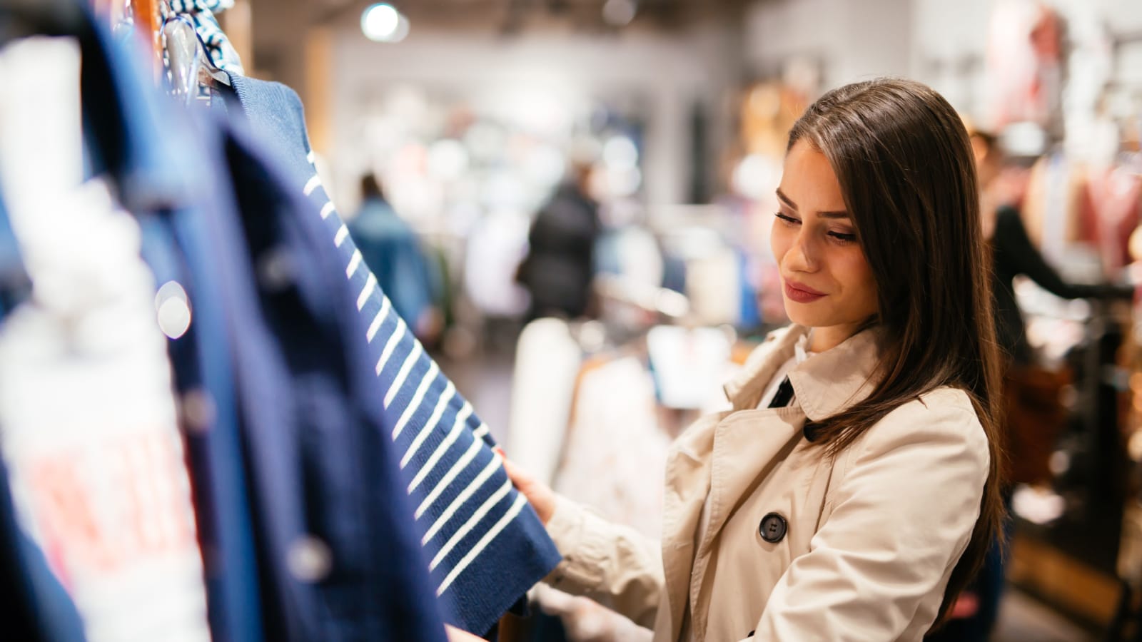 A woman shopping for clothes in a retail store. She admires a blue and white stripped top on the hanger.