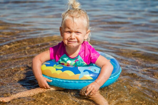 Baby girl crying, sitting on the sand at the beach near the ocean.Toddler is unhappy and having tantrum