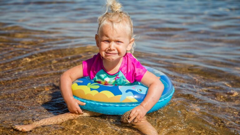 Baby girl crying, sitting on the sand at the beach near the ocean.Toddler is unhappy and having tantrum