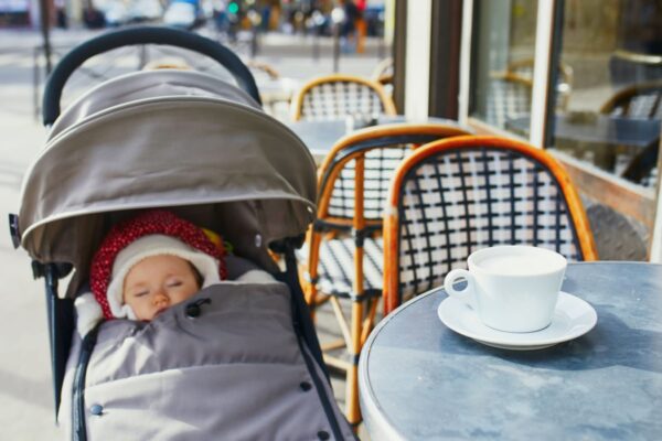 Baby girl sleeping in pram on outdoor terrace of Parisian street cafe with cup of hot coffee on the table. Going out with kids