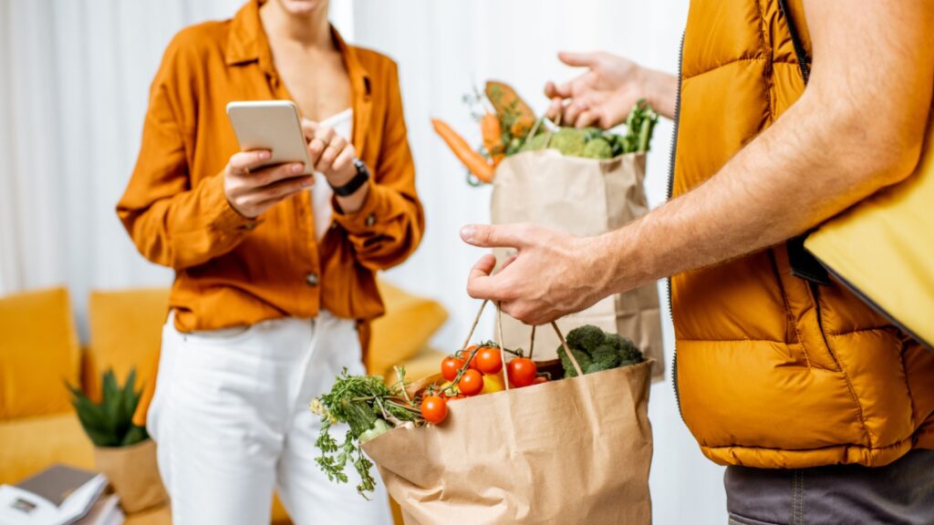 Courier in uniform with thermal bag delivering fresh groceries in paper bags to a client home. Woman with smartphone checking her order