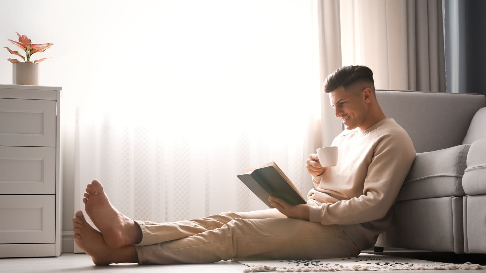 Man with cup of hot drink reading book at home. Floor heating concept