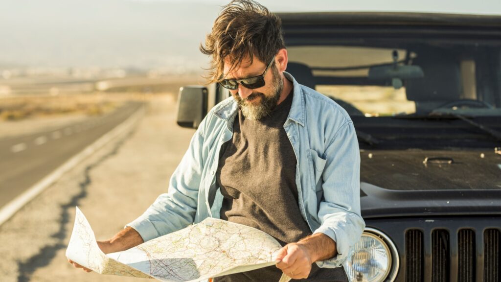 Man looking at map leaning on vehicle at roadside. Mature man checking location of destination on paper map standing outside car. Man searching for navigational route using paper map at roadside