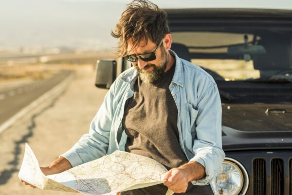 Man looking at map leaning on vehicle at roadside. Mature man checking location of destination on paper map standing outside car. Man searching for navigational route using paper map at roadside