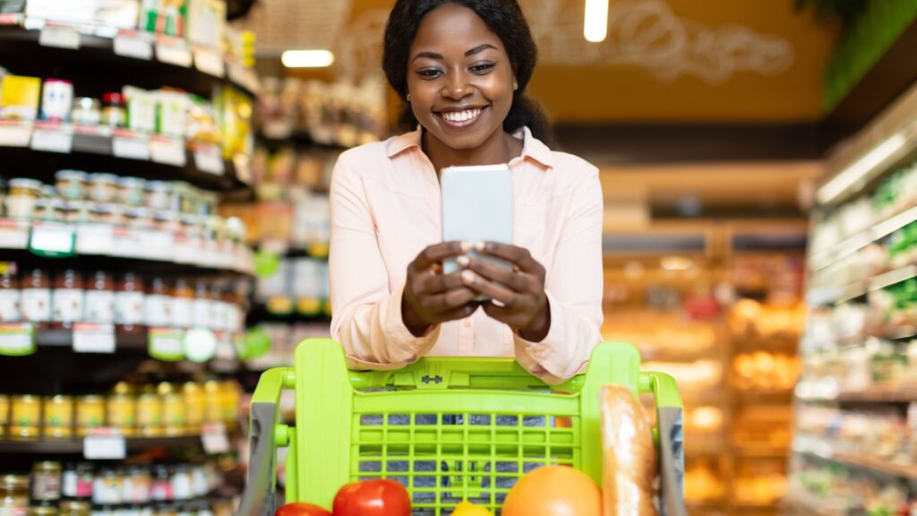 Happy African American Lady Doing Grocery Shopping On Mobile Phone Via Application, Posing With Cellphone And Cart In Supermarket. Woman