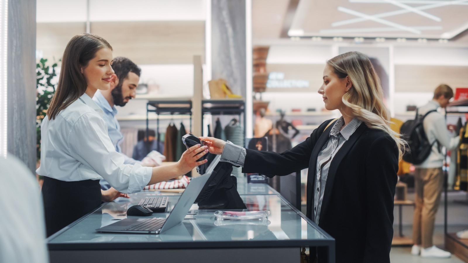 Clothing Store Checkout Cashier Counter: Beautiful Young Woman Buys Blouse from Friendly Retail Sales assistant, Paying with Contactless Credit Card.