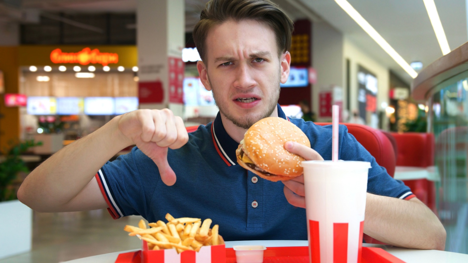 Young negative sad angry unpleased dissatisfied man guy is sitting on food court holding in hands burger hamburger showing thumb down dislike gesture. Fast junk unhealthy lifestyle. Stop fast food