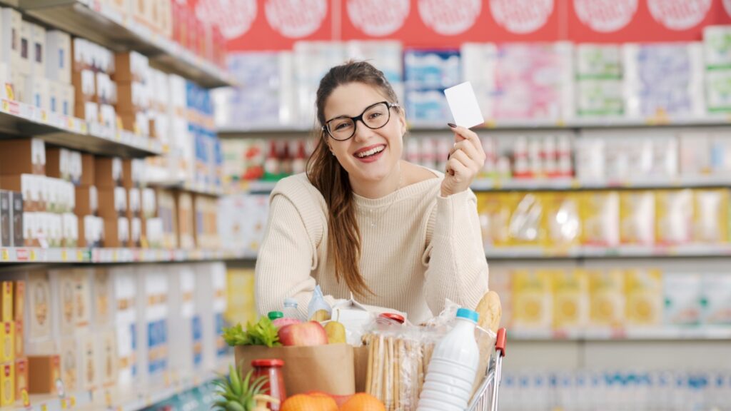 Smiling customer leaning on a full shopping cart and showing a blank loyalty card at the supermarket, grocery loyalty program and discounts concept