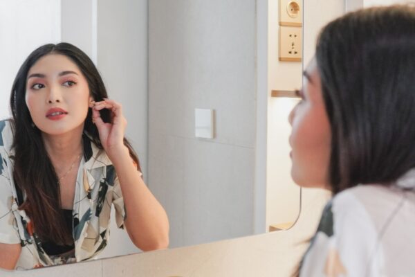 Beautiful young Asian woman wearing patterned shirt looking at herself in the mirror while putting on earring. Beauty concept.