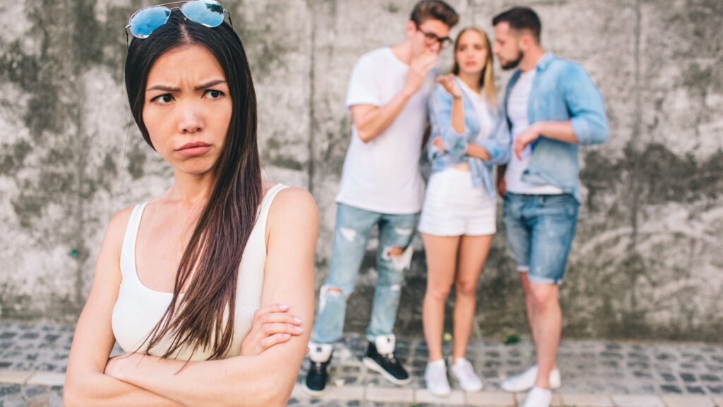 Upset young chinese girl is standing in front of picture and looking to theside. She has crossed her hands. Her friends are gossiping behind her back. They are standing on grey background.