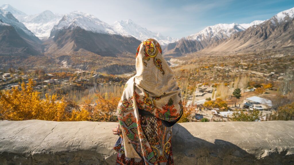 A woman wearing traditional dress sitting on wall and looking at Hunza valley in autumn season, Gilgit Baltistan in Pakistan, Asia