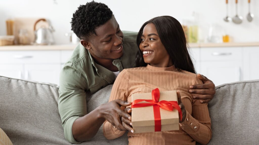 Loving african american man hugging his wife, giving gift box, making great surprise for St. Valentines Day, home interior, free space. Happy black guy embracing his girlfriend, holding present