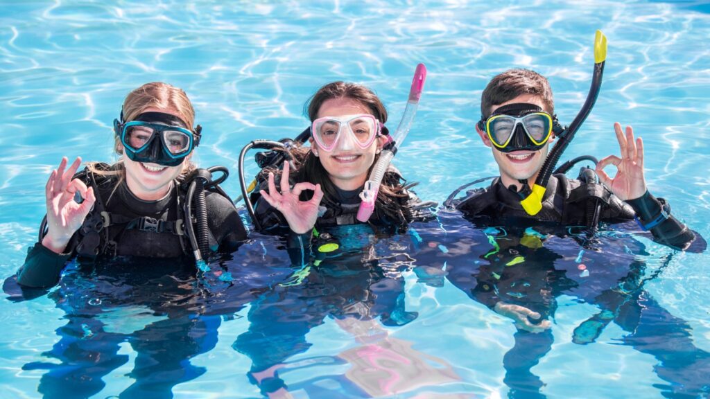 A group of happy scuba divers smiling at the camera with their dive masks and gear on in a pool showing the OK sign