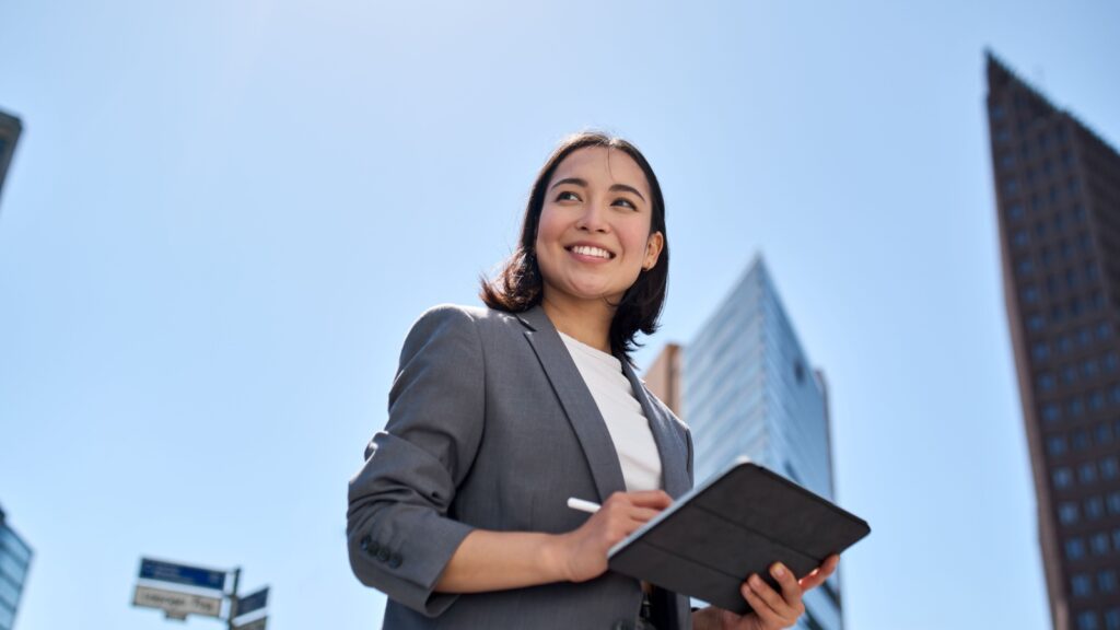 Smiling young Asian business woman leader entrepreneur, professional manager holding digital tablet computer using software applications standing on the street in big city on sky background.