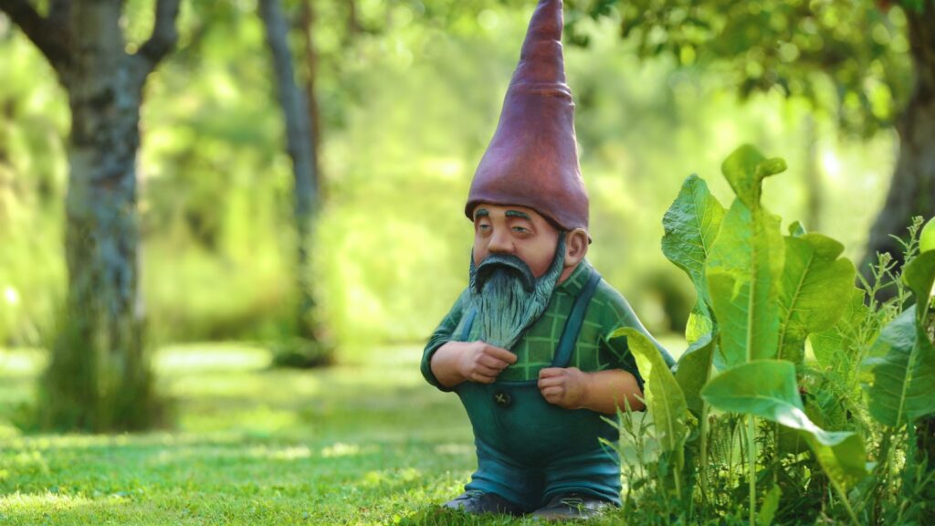 Garden dwarf with a long beard and a fairy cap in a beautiful summer garden during midday. A fairy-tale character made of cement and painted in bright colors. Portrait of garden gnome.