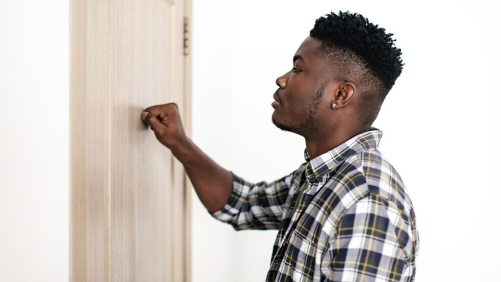 Black Millennial Man Knocking At Closed Door Of Apartment Indoors. Man Standing Near Entry Of Flat Posing In Doorway. Real Estate Business And Offers Concept. Side View Shot