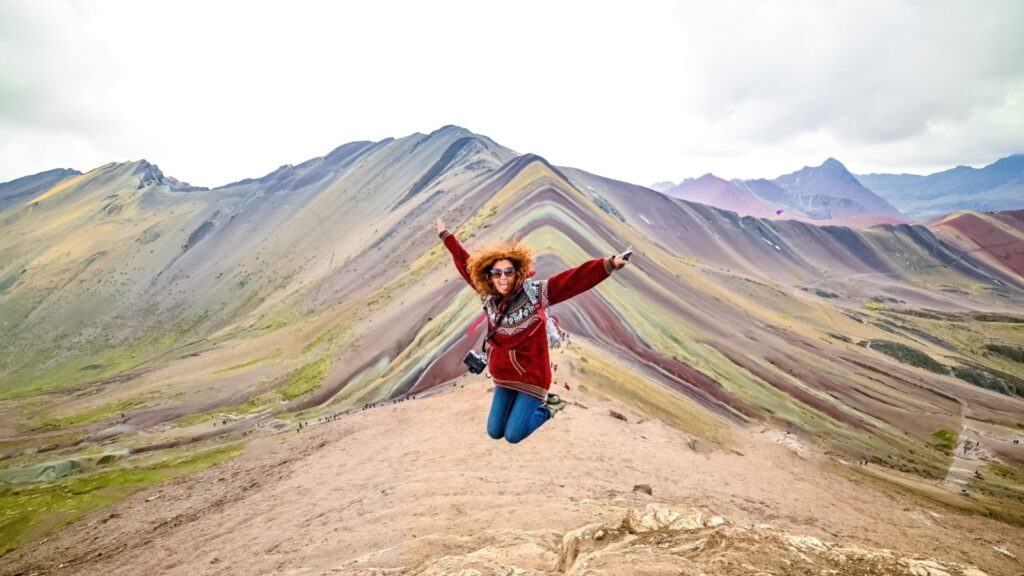 Young red haired smiling girl jumping in front of the Vinicunca Rainbow Mountain, Peru