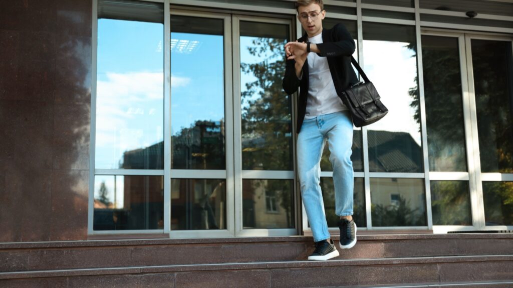 Emotional young man checking time while going down stairs near modern building outdoors. Being late