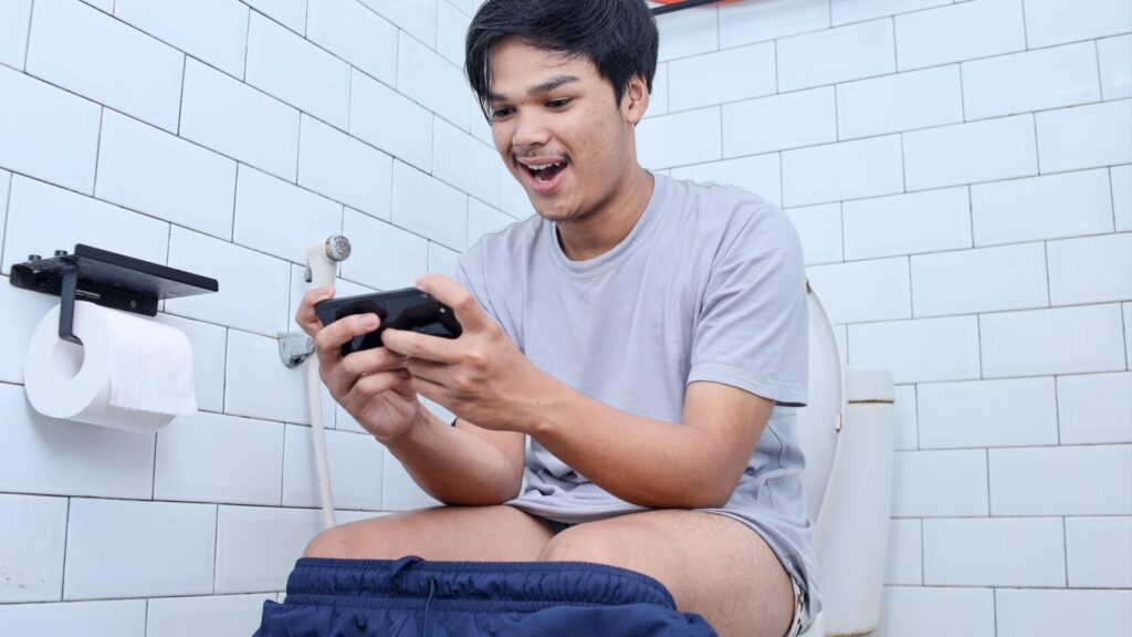 Happy Asian young man using mobile phone while sitting on toilet bowl in house bathroom playing online games